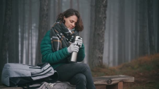 Mujer joven bebe té caliente de un termo mientras está sentada en un banco en un bosque — Vídeo de stock
