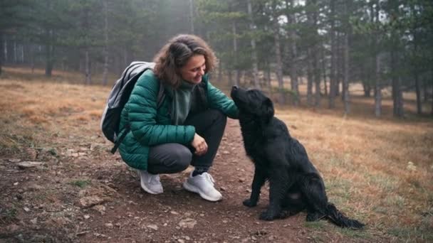 Mujer joven acariciando a su lindo perro negro mientras la pasea en el bosque de otoño — Vídeos de Stock