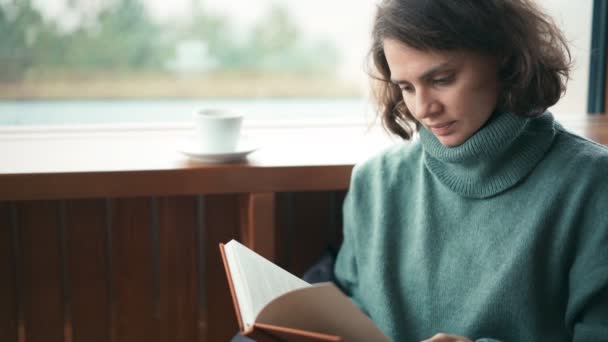 Una joven leyendo un libro y tomando café de una taza en el interior. — Vídeos de Stock