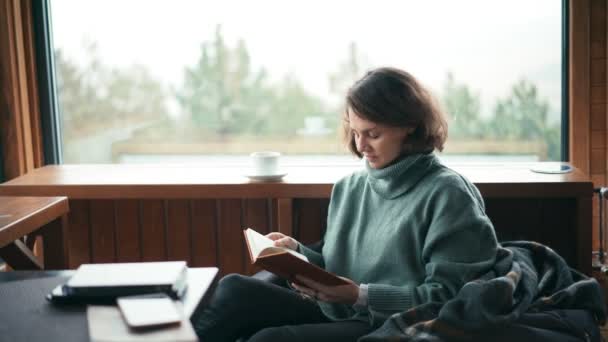 Een jonge vrouw in haar woonkamer die een boek leest en koffie drinkt — Stockvideo