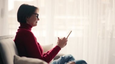 Kanepede otururken akıllı telefonunu kullanan gözlüklü rahat genç bir kadın..