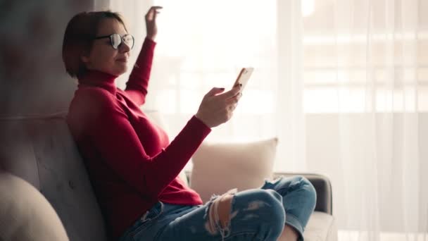 Jeune femme détendue dans des lunettes à l'aide de son smartphone assis sur le canapé. — Video