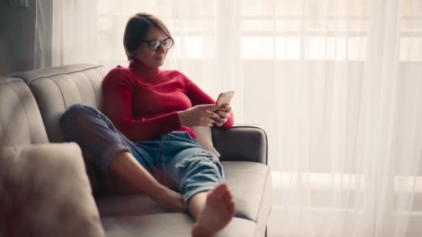 Zrelaksowana młoda kobieta w okularach za pomocą smartfona siedząc na kanapie. — Wideo stockowe