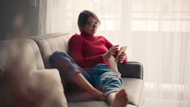 Zrelaksowana młoda kobieta w okularach za pomocą smartfona siedząc na kanapie. — Wideo stockowe