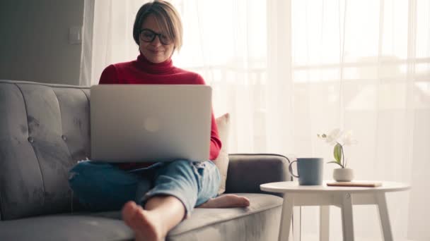 Mladá žena pracuje na svém notebooku a pije kávu, zatímco sedí na gauči — Stock video