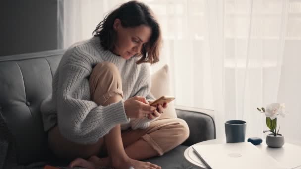 Kanepede otururken akıllı telefon kullanan rahat genç bir kadın. — Stok video