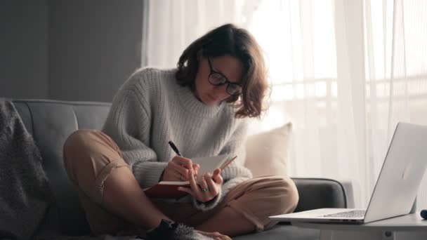 Mladá žena se dívá na monitor notebooku a dělá si poznámky do svého notebooku — Stock video