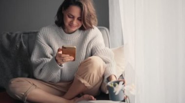 Akıllı telefon kullanan ve fincandan kahve içen rahat genç bir kadın.