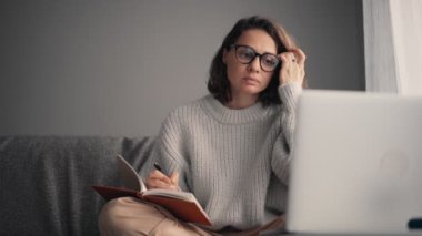 Genç bir kadın dizüstü bilgisayara bakıyor ve not defterine not alıyor.