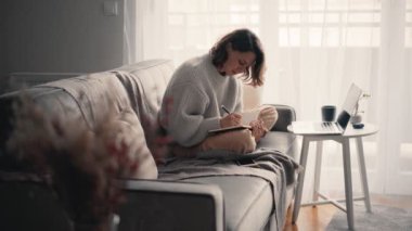 Genç bir kadın dizüstü bilgisayara bakıyor ve not defterine not alıyor.