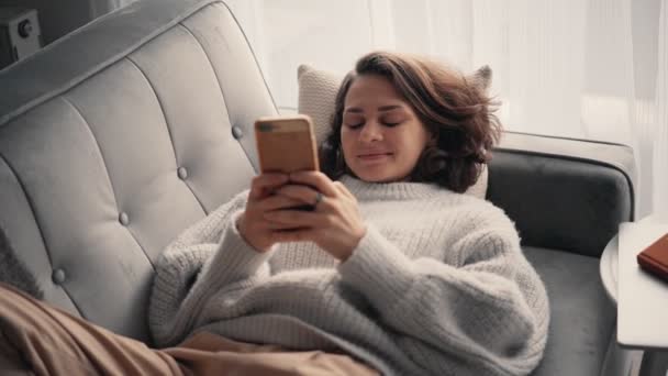 Uma jovem relaxada usando seu smartphone enquanto estava deitada no sofá. — Vídeo de Stock