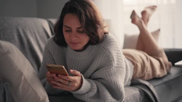 Uma jovem relaxada usando seu smartphone enquanto estava deitada no sofá. — Vídeo de Stock