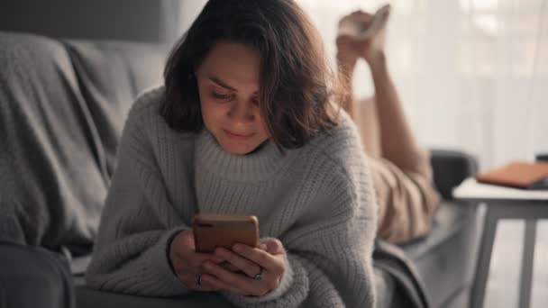 Uvolněná mladá žena pomocí svého smartphonu, zatímco leží na pohovce. — Stock video