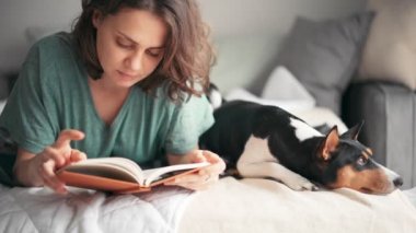 Genç bir kadın, Basenji köpeğiyle yatakta yatarken kitap okuyor.