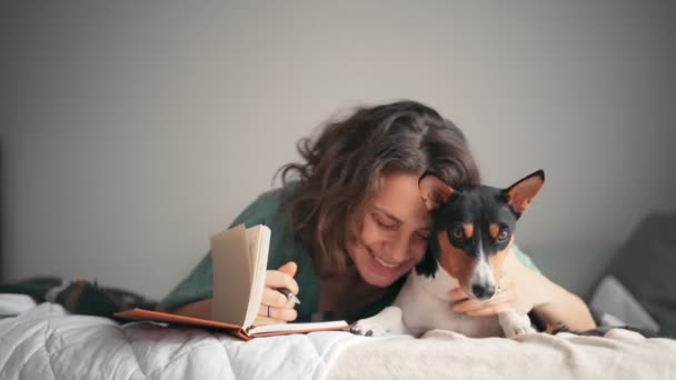 Egy fiatal nő naplót ír, miközben az ágyban fekszik a basenji kutyájával. — Stock videók