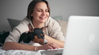 Bir kadın, köpeğiyle yatakta yatarken laptopuyla video görüşmesi yapıyor.