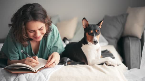 Молодая женщина пишет дневник, лежа в постели со своей собакой. — стоковое видео