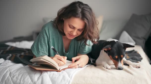 Une jeune femme écrit un journal alors qu'elle est couchée au lit avec son chien basenji — Video