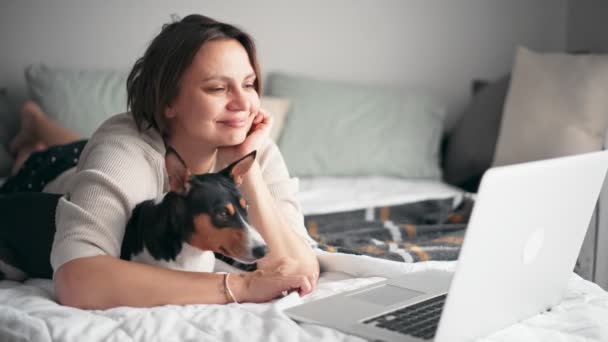 Una mujer de mediana edad está trabajando en su computadora portátil mientras está acostada en la cama con su perro — Vídeo de stock