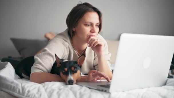 Une femme d'âge moyen travaille sur son ordinateur portable alors qu'elle est allongée sur le lit avec son chien — Video