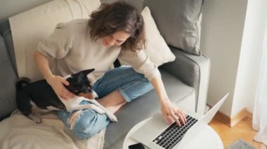 Genç bir kadın tatlı köpeğiyle kanepede otururken dizüstü bilgisayarda çalışıyor.