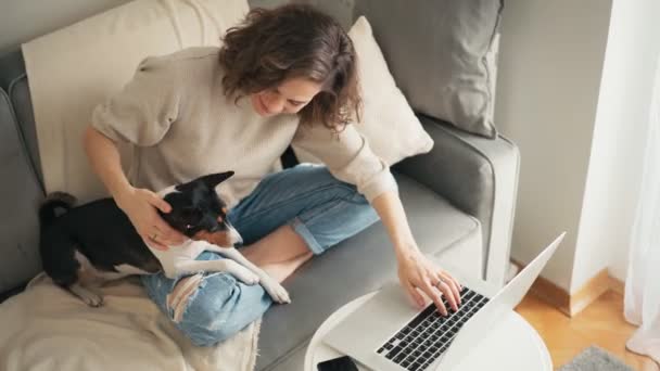 Une jeune femme travaillant sur un ordinateur portable assis sur un canapé avec son chien mignon — Video