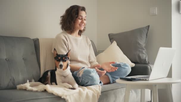 Женщина принимает видеозвонок на ноутбук, сидя на диване со своей собакой — стоковое видео