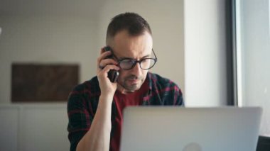 Yakışıklı, ciddi bir adam evde dizüstü bilgisayarla konuşurken telefonda konuşuyor.