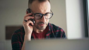 Yakışıklı, ciddi bir adam evde dizüstü bilgisayarla konuşurken telefonda konuşuyor.