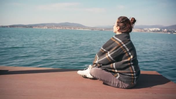 Joven hermosa mujer se envuelve en cuadros mientras se sienta en un muelle junto al mar — Vídeo de stock