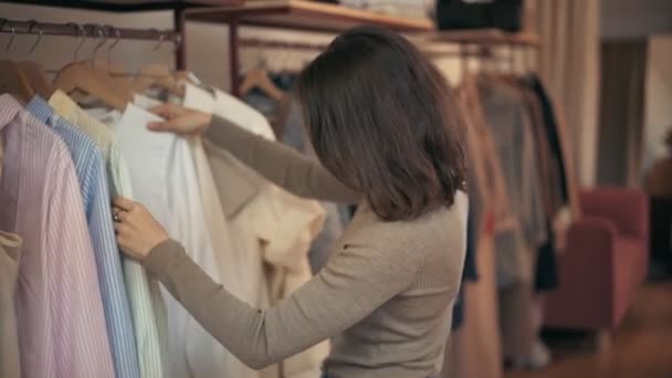 Una giovane donna esamina i vestiti sulle grucce nel negozio — Video Stock