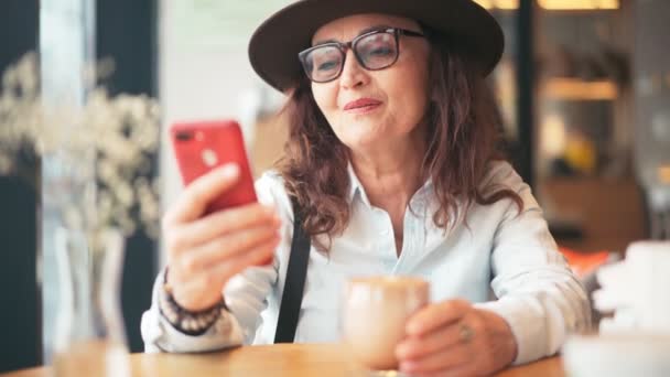 Een volwassen stijlvolle vrouw met hoed en bril het nemen van een video gesprek op een smartphone — Stockvideo