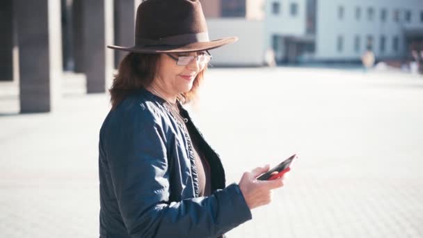A senior woman in glasses and a hat using her smartphone — Vídeo de Stock