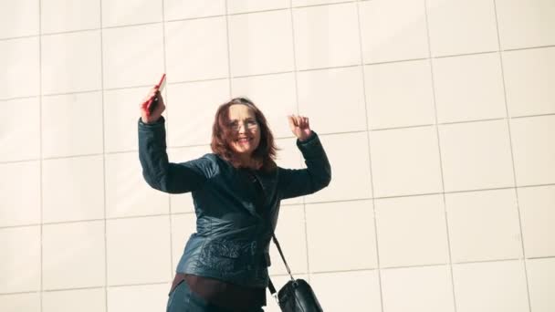 Een dolblij senior vrouw met een bril dansend op straat op een zonnige dag — Stockvideo