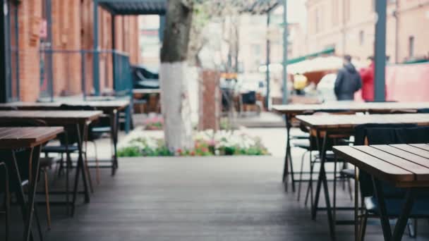 Fából készült asztalok az étterem üres utcai verandáján, emberek mennek el mellette. — Stock videók