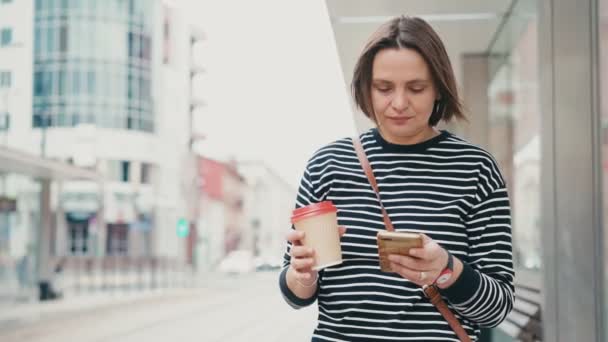 Eine junge erwachsene Frau blickt auf den Smartphone-Bildschirm, während sie auf einen Bus wartet — Stockvideo