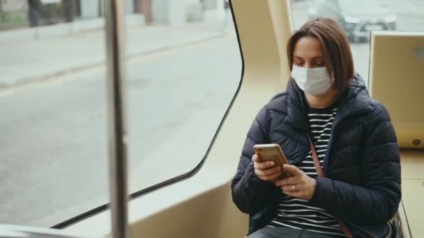 Молода доросла жінка в масці для обличчя використовує смартфон під час їзди на автобусі — стокове відео