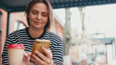 Genç yetişkin bir kadın açık bir verandada oturur ve akıllı telefonunu kullanır.