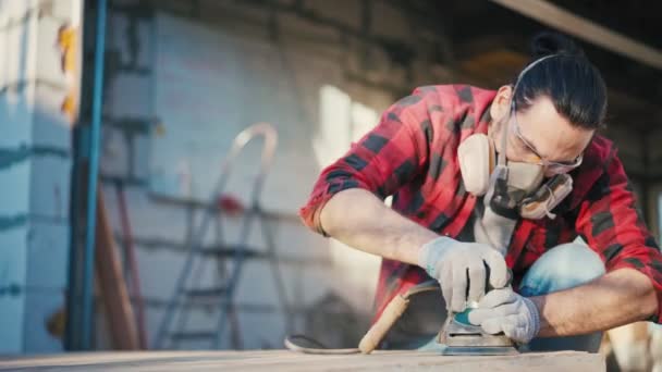 Een jongeman in een bouwmasker slijpt een parketplank met een schuurmachine — Stockvideo