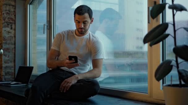 Um homem bonito senta-se em um peitoril largo da janela e aprecia a vista da cidade da noite — Vídeo de Stock