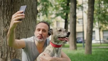 Yakışıklı sakallı bir adam sevimli gri köpeğini kucaklıyor ve selfie çekiyor.