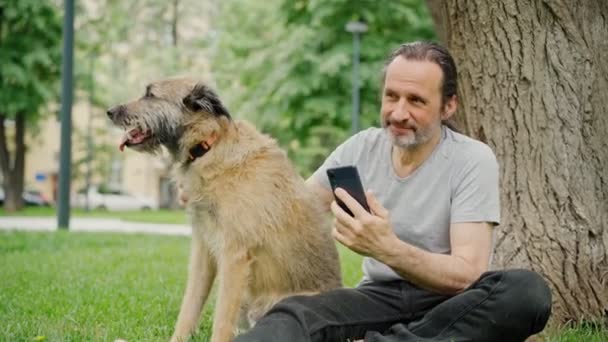 Een knappe man met een baard zit op het gras in het park en streelt zijn hond — Stockvideo