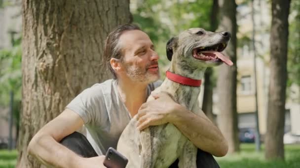 Een knappe man zit op het gras in het park en knuffelt zijn hond. — Stockvideo