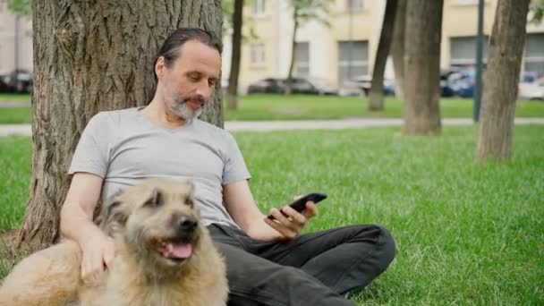 Een knappe man met een baard zit op het gras in het park en streelt zijn hond — Stockvideo