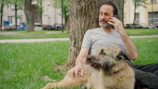 Un hombre se sienta en el césped del parque acariciando a su perro y hablando por teléfono — Vídeos de Stock