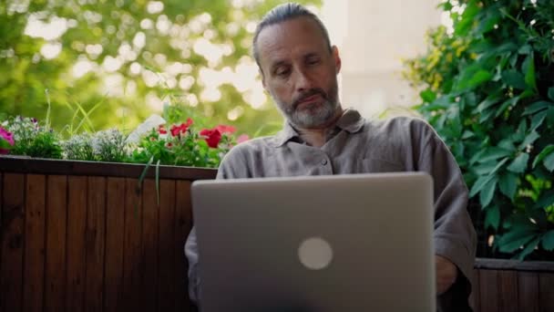 Um belo homem adulto médio digitando em um laptop enquanto sentado no terraço aberto — Vídeo de Stock