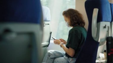 Trene binerken akıllı telefonunu kullanan maskeli genç bir kadın..