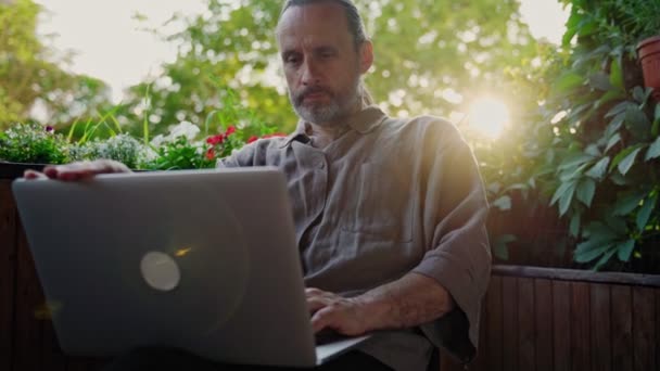 En stilig vuxen man som skriver på en bärbar dator medan han sitter på den öppna terrassen — Stockvideo