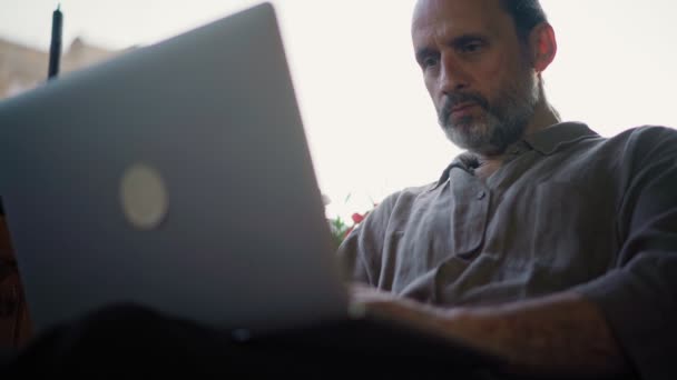 Um belo homem adulto médio digitando em um laptop enquanto sentado no terraço aberto — Vídeo de Stock