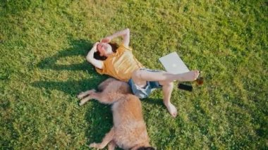 A young woman relaxing by lying on a lawn with her quite shaggy dog.